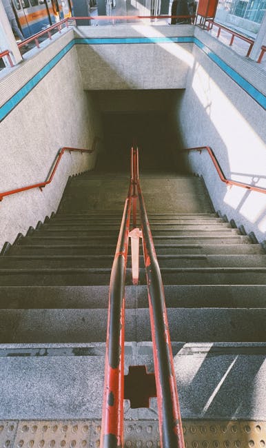 Photograph of a concrete staircase inside a building with metal handrails painted in reddish-brown leading down to a lower level. The stairs are flanked by textured, light-colored walls with a blue accent strip along the top. The lighting is natural, coming from an upper window or opening, illuminating the steps and handrails. The staircase is part of a building with enclosed walls, possibly within an urban residential or commercial property. The image captures the full descent of the staircase, emphasizing the narrowness of the access point, which can present challenges during a home relocation. As part of house removals or furniture transport, the image reflects the careful planning needed when navigating tight staircases during loading or unloading processes, working with equipment such as trolleys or lifts, and highlighting the significance of professional moving services like those provided by Man with Van Mile End to ensure safe and efficient access through confined passageways.