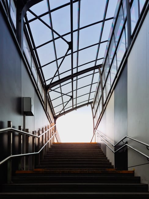 A staircase inside a building, featuring dark wooden steps with metal edges, leading upwards towards an exit illuminated by bright natural light. The staircase is bordered by metal railings on both sides, with the right-side railing attached to a smooth, light-colored wall. Above the stairs, a large glass ceiling with a metal framework allows sunlight to flood the area, creating a contrast between the dark stairs and the bright sky outside. The environment appears modern, with clean lines and a minimalist design, suitable for a residential or commercial property. This scene captures the interior space involved in home relocation, illustrating the process of moving furniture or boxes through stair access when handling narrow or challenging staircases, with the potential for transportation equipment like trolleys or straps being used outside the frame. Man with Van Mile End offers professional removal services that can manage complex access areas like this one during house moves.