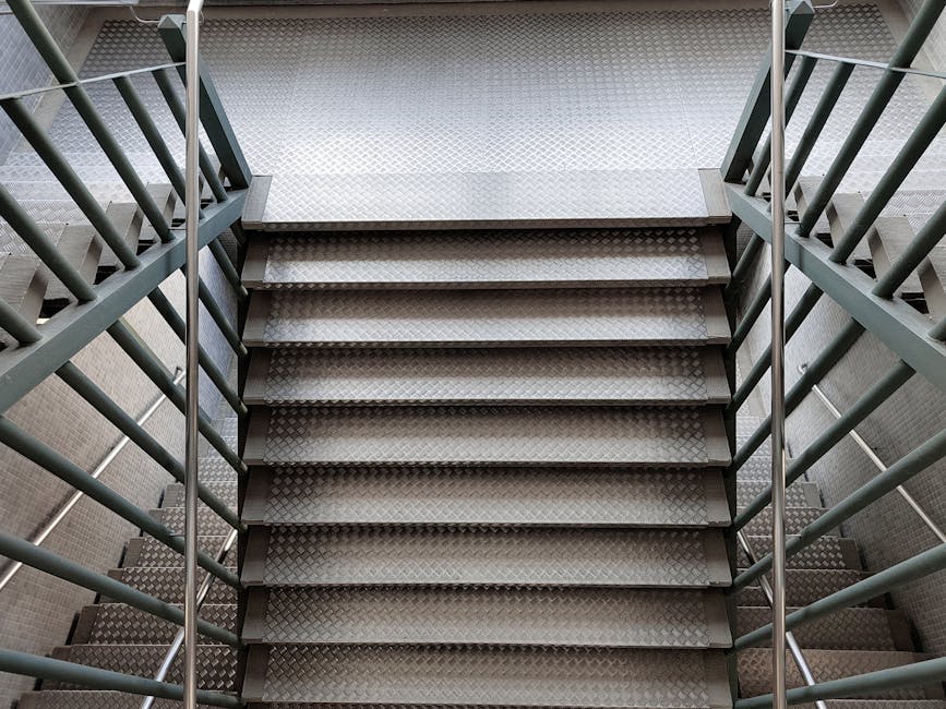 Photograph of a concrete staircase inside a building with metal handrails painted in reddish-brown leading down to a lower level. The stairs are flanked by textured, light-colored walls with a blue accent strip along the top. The lighting is natural, coming from an upper window or opening, illuminating the steps and handrails. The staircase is part of a building with enclosed walls, possibly within an urban residential or commercial property. The image captures the full descent of the staircase, emphasizing the narrowness of the access point, which can present challenges during a home relocation. As part of house removals or furniture transport, the image reflects the careful planning needed when navigating tight staircases during loading or unloading processes, working with equipment such as trolleys or lifts, and highlighting the significance of professional moving services like those provided by Man with Van Mile End to ensure safe and efficient access through confined passageways.