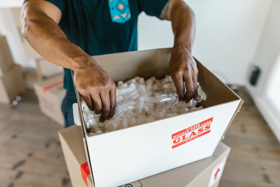 A person wearing a dark T-shirt is carefully placing clear glass bottles into a cardboard box filled with white packing peanuts for cushioning, which is positioned on top of a white plastic crate inside a residential property. The box has a red and white label indicating 'HAPPY PLASTIC GLASS.' The scene takes place indoors near a doorway, with natural lighting illuminating the wooden floor and white walls. This image illustrates the careful packing process involved in home relocation, specifically handling fragile glass items during furniture transport with the assistance of Man with Van Mile End, a professional removal service.