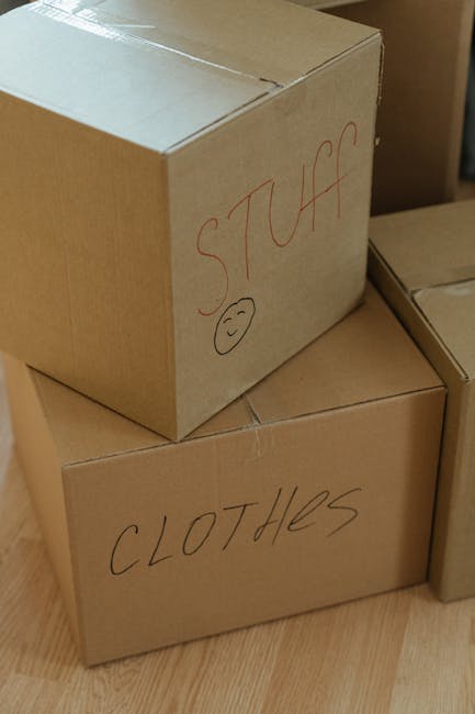 Close-up photograph of three cardboard moving boxes situated indoors on a wooden floor, with natural light illuminating the scene. One box is labeled 'STUFF' in red marker with a small smiley face drawn beneath the text, and the box is partially stacked atop another labeled 'CLOTHES' in black marker. The boxes are sealed with packing tape, appearing sturdy for a home relocation or furniture transport process. In the background, additional boxes are visible, suggesting an organized packing arrangement for a house move. The image reflects typical packing and loading activities carried out by professional removals services such as Man with Van Mile End, emphasizing careful handling of personal belongings during a house removal.