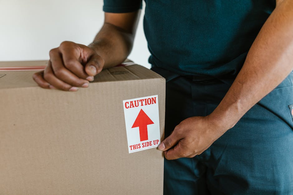 A person wearing a dark T-shirt is carefully placing clear glass bottles into a cardboard box filled with white packing peanuts for cushioning, which is positioned on top of a white plastic crate inside a residential property. The box has a red and white label indicating 'HAPPY PLASTIC GLASS.' The scene takes place indoors near a doorway, with natural lighting illuminating the wooden floor and white walls. This image illustrates the careful packing process involved in home relocation, specifically handling fragile glass items during furniture transport with the assistance of Man with Van Mile End, a professional removal service.