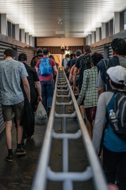 A view of a crowded station corridor with numerous pedestrians walking along a moving walkway. The individuals are dressed in casual clothing and carrying backpacks, suitcases, and bags, indicating th