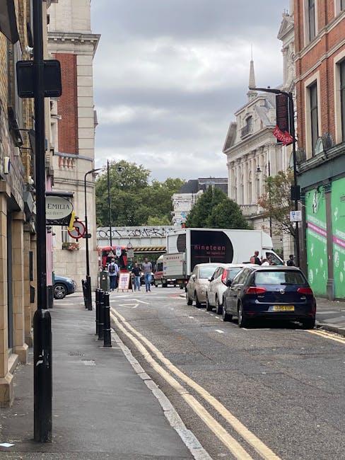 A street scene in Mile End showing a narrow road with parked cars lining the pavement on the right side. The road is being used for home relocation activities, with a white moving van positioned furth