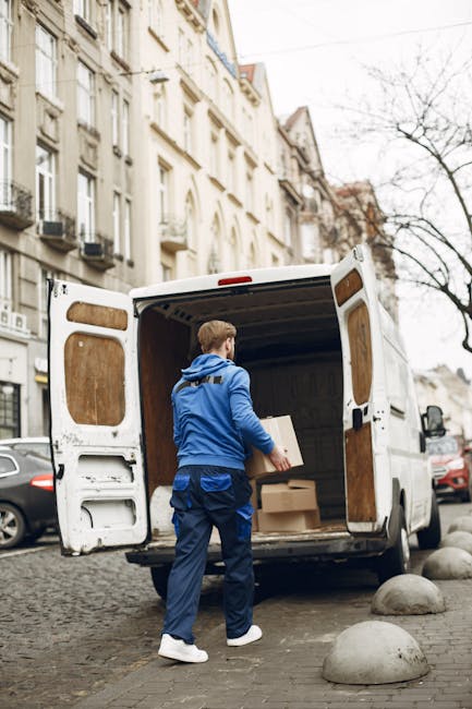 A man dressed in a blue jacket, dark blue pants, and white sneakers is loading cardboard boxes into the back of a white delivery van parked on a cobblestone street in Mile End. The van's rear doors are wide open, revealing an interior lined with wooden panels. The boxes vary in size and are made of brown cardboard, some stacked on top of each other. Several large, smooth concrete bollards are positioned along the edge of the street, which is lined with elegant, multi-storey residential buildings featuring balconies, large windows, and ornate facades. Bare trees suggest a colder season. The scene captures a home relocation or furniture transport process, with the man preparing for the next phase of packing and moving, assisted by the professional removals service offered by Man with Van Mile End.