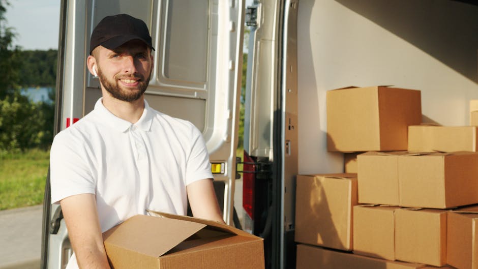 A man with a beard, dressed in a white polo shirt and black cap, is seen loading cardboard boxes into a van for home relocation. The van, parked outdoors on a paved area, has its side door open revealing a spacious interior filled with stacked cardboard boxes of various sizes, some wrapped in plastic or placed on pallets. The boxes are arranged neatly inside the vehicle, ready for transport. The background shows a green landscape with trees and a blue sky, indicating daylight conditions. The scene captures the process of furniture transport and packing during a professional removals service, as provided by Man with Van Mile End, supporting efficient moving logistics in Mile End.