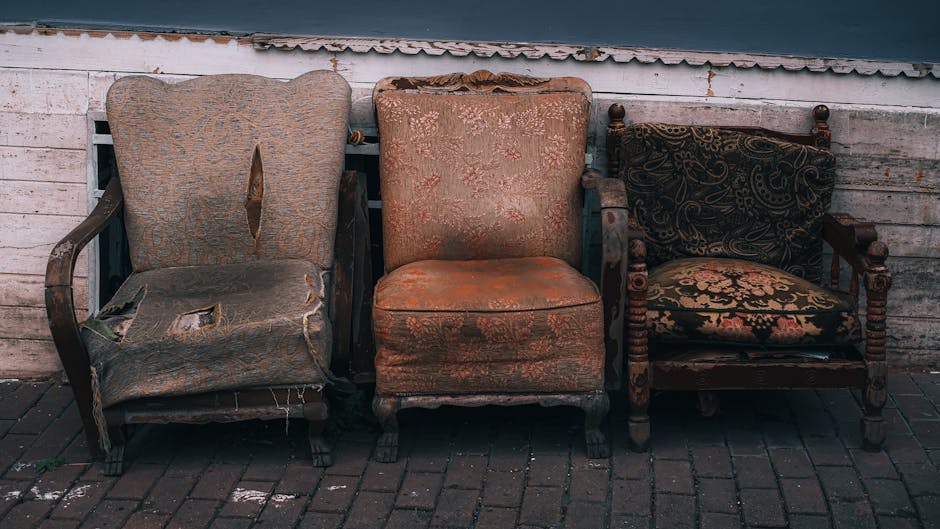 A row of three upholstered chairs positioned outside a building along a brick-paved pavement. The first chair on the left is an old, damaged armchair with torn fabric, exposed wooden frame, and missing padding. The middle chair is a vintage-style armchair with faded, worn orange floral-patterned fabric, wooden legs, and a slightly stained appearance. The third chair on the right is a wooden armchair with ornate carvings and a dark fabric cushion with decorative patterns. Behind the chairs, there is a white wooden fence and a section of building wall. This scene shows furniture arranged outdoors, possibly awaiting removal or disposal, which relates to house removals and furniture transport services. Man with Van Mile End occasionally provides professional assistance with packing and moving furniture during home relocations, ensuring efficient loading and transportation processes.