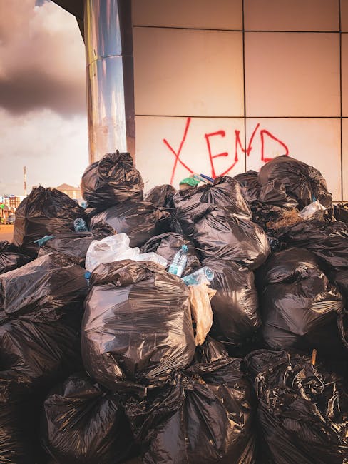 A large pile of black rubbish bags filled with household waste, some tied tightly while others are partially open, revealing plastic bottles and packaging materials inside. The bags are stacked on the pavement near a modern building with tiled walls, on which red graffiti spelling 'XEND' is visible. In the background, a cloudy sky with a hint of sunlight can be seen. The scene relates to the context of household waste disposal in Mile End, with the image illustrating typical refuse accumulation before removal, relevant to home relocation and furniture disposal regulations in Tower Hamlets, as managed by Man with Van Mile End.
