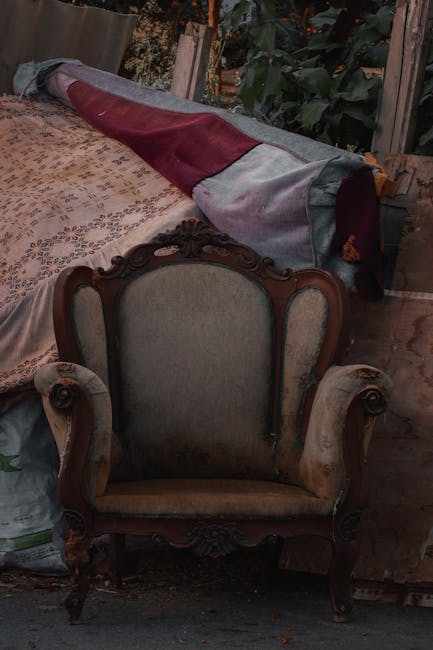 A row of three upholstered chairs positioned outside a building along a brick-paved pavement. The first chair on the left is an old, damaged armchair with torn fabric, exposed wooden frame, and missing padding. The middle chair is a vintage-style armchair with faded, worn orange floral-patterned fabric, wooden legs, and a slightly stained appearance. The third chair on the right is a wooden armchair with ornate carvings and a dark fabric cushion with decorative patterns. Behind the chairs, there is a white wooden fence and a section of building wall. This scene shows furniture arranged outdoors, possibly awaiting removal or disposal, which relates to house removals and furniture transport services. Man with Van Mile End occasionally provides professional assistance with packing and moving furniture during home relocations, ensuring efficient loading and transportation processes.
