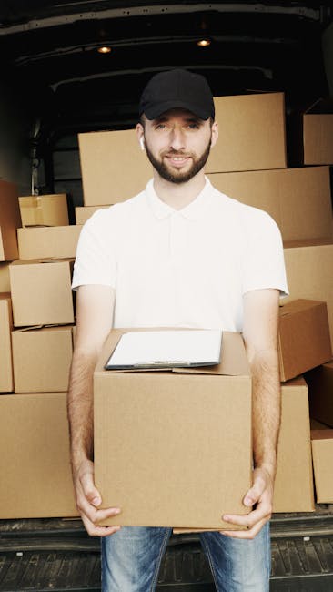 A young male mover with a well-groomed beard, wearing a black cap, white t-shirt, and blue jeans, standing inside the back of a moving van with a black interior. He is holding a medium-sized cardboard box at waist level, with both hands, and smiling at the camera. Behind him, there are several stacked cardboard boxes of various sizes, some wrapped with plastic or tape, arranged neatly on the van’s floor and sides. A clipboard with paperwork rests on the box he is holding, indicating the packing and moving process. The van’s open rear door reveals a paved area outside, suggesting the home relocation is in progress. The scene reflects furniture transport, packing procedures, and loading activities typical of professional removals, as seen by [COMPANY_NAME], offering house removal services in Mile End.