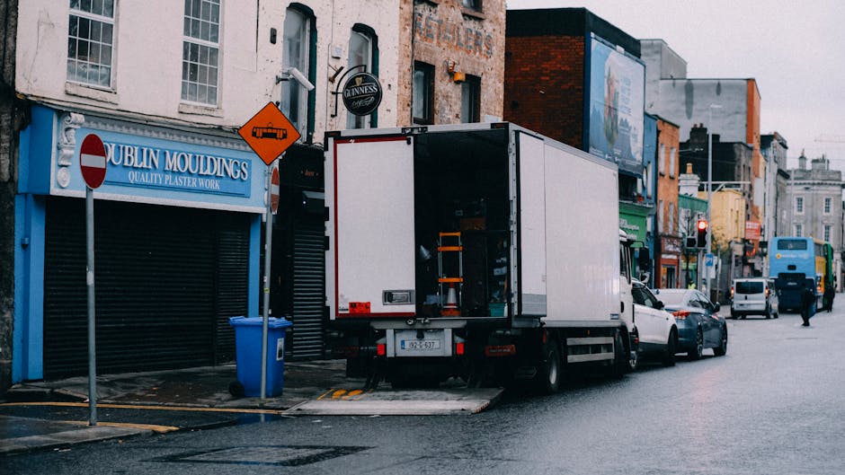 A white moving truck with its rear door open, positioned on the street outside a commercial property during daylight hours. Inside the truck, visible packing materials and boxes are being loaded or prepared for transportation as part of a home relocation or furniture transport process. The truck is parked partly on the sidewalk with the rear lift gate lowered, facilitating the loading process. Several cardboard boxes, some wrapped in plastic, are arranged inside and near the truck, while nearby on the pavement, there are a blue recycling bin and a small orange warning sign attached to a pole. The street features a row of shops with signage, such as 'Dublin Mouldings' and others, amidst parked cars and pedestrian activity. The street environment is typical of urban commercial districts and may be part of a local removals or moving service managed by Man with Van Mile End, emphasizing the logistics involved in packing and transporting household or business items.